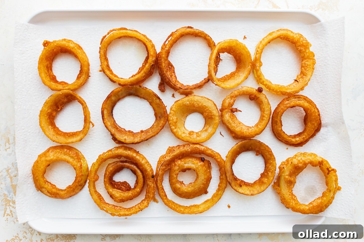 Crispy Gluten-Free Onion Rings 12 Freshly fried, golden gluten-free onion rings neatly draining on paper towels on a baking pan, absorbing excess oil.