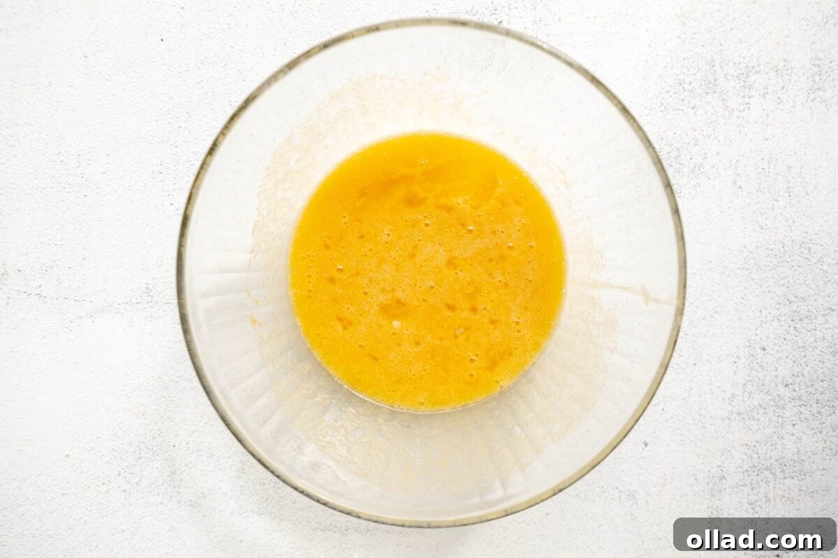 A clear glass bowl containing a mixture of wet ingredients for almond flour blueberry muffins, with a smooth golden color against a light countertop background.