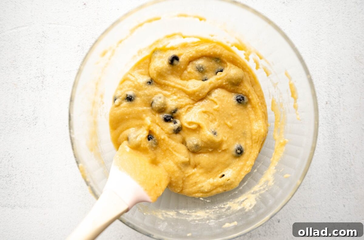 A clear glass bowl with almond flour muffin batter folded together with fresh blueberries, a white spatula resting inside the bowl.