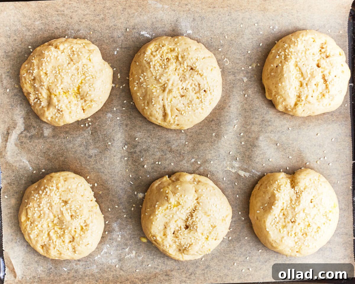 risen and shaped gluten-free hamburger bun dough topped with sesame seeds on a baking sheet.