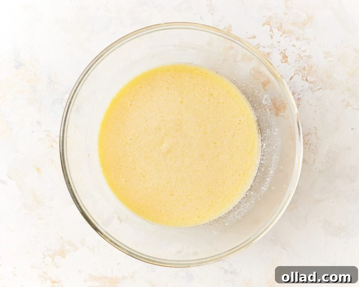Irresistible Gluten-Free Muffins 4 A large glass mixing bowl filled with a smooth, pale yellow liquid mixture, showing the perfectly combined wet ingredients (eggs, buttermilk, sour cream, melted butter, oil, and vanilla extract) for gluten-free muffins.