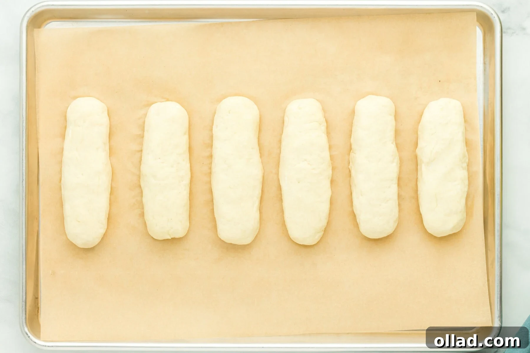 Six perfectly shaped gluten-free hot dog bun logs arranged on a parchment-lined baking sheet, ready for proofing.