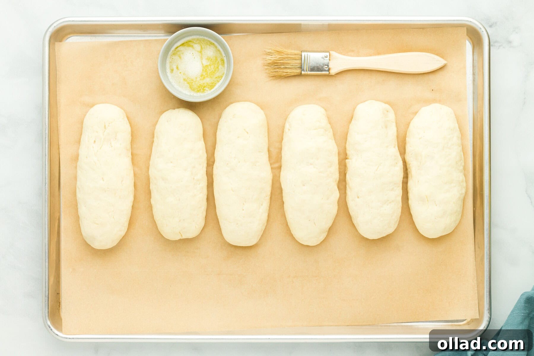Proofed gluten-free hot dog bun dough logs on a baking sheet, alongside a bowl of melted butter and a basting brush.