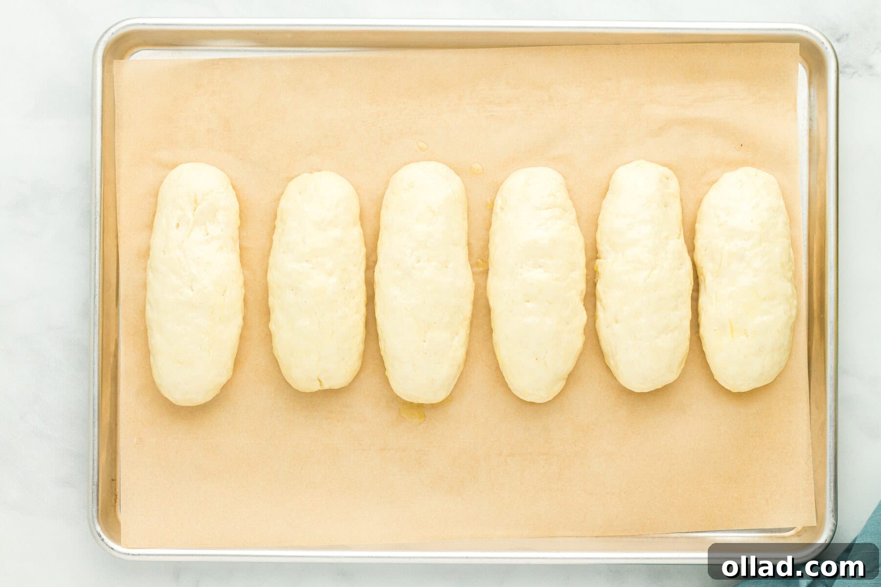 Gluten-free hot dog bun dough, now fully proofed and expanded, on a parchment-lined baking sheet, brushed with butter.