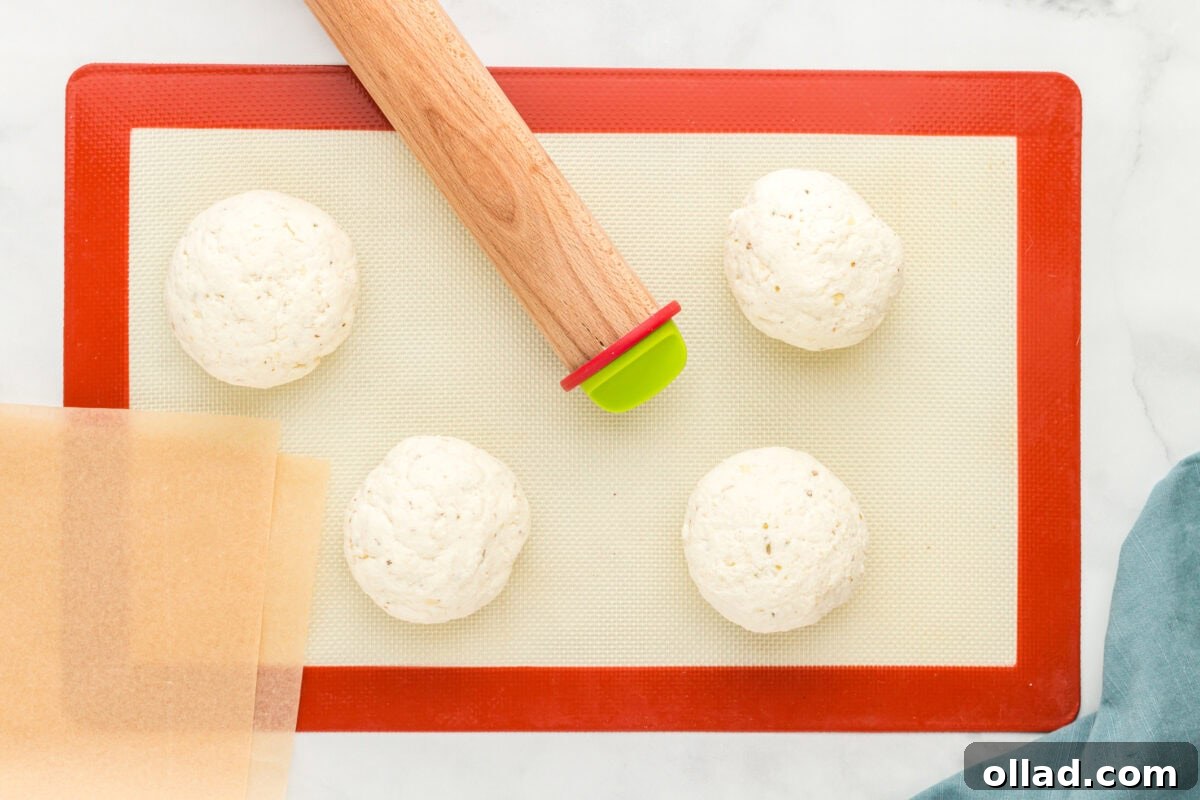 Pillowy Gluten-Free Flatbread 6 Four evenly shaped gluten-free flatbread dough balls placed on a red-edged silicone baking mat with sheets of parchment paper and a wooden rolling pin nearby, on a white marble surface with a blue cloth napkin.
