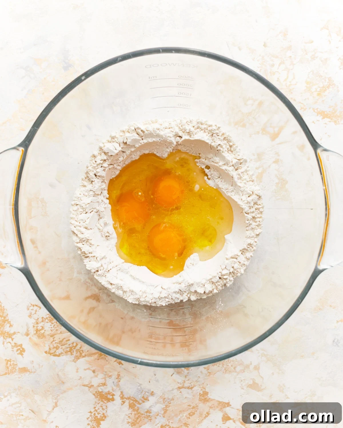 Eggs, water, and olive oil in the center of a flour well in a glass bowl.