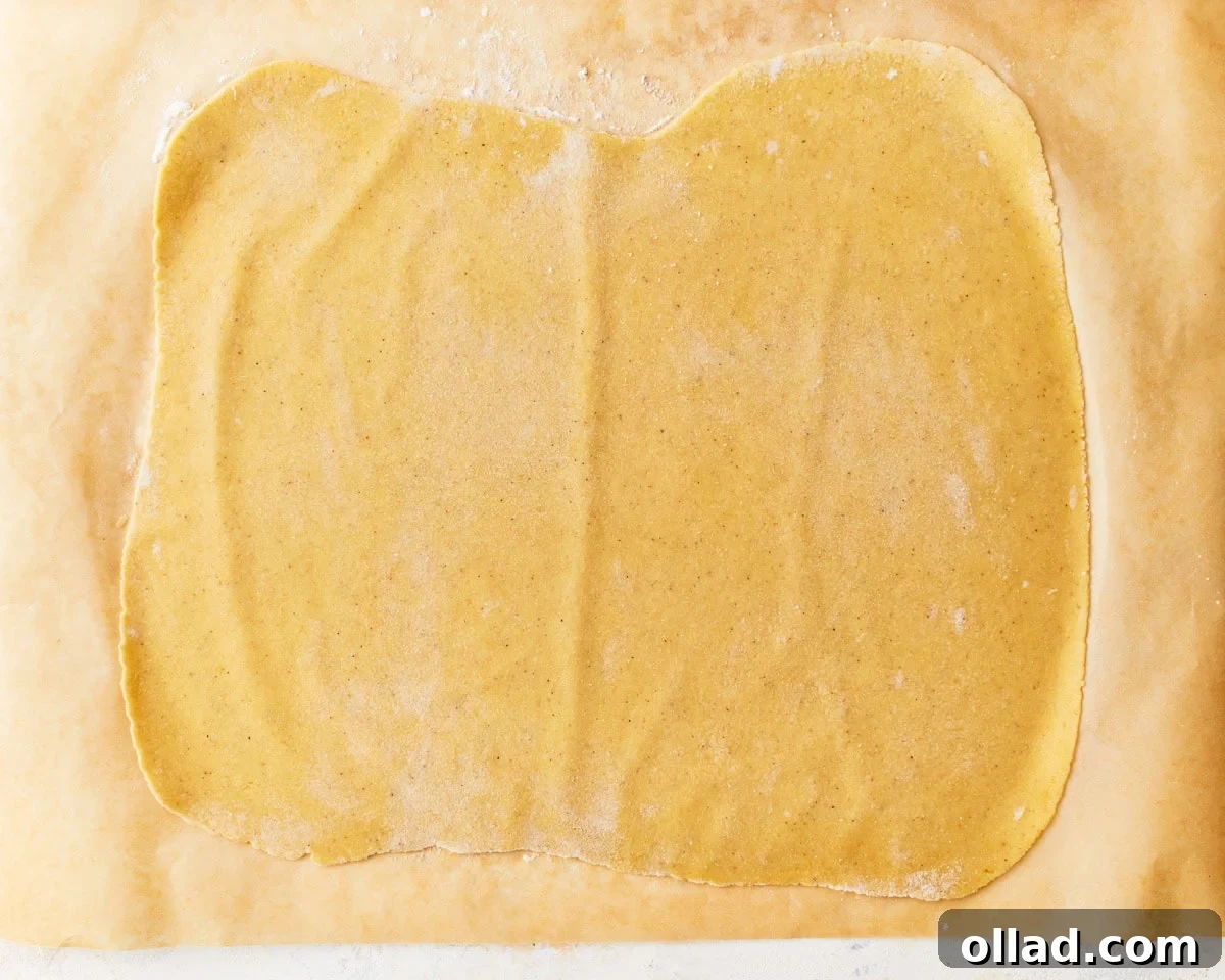 A sheet of gluten-free egg noodle dough expertly rolled out thinly on parchment paper with a rolling pin.