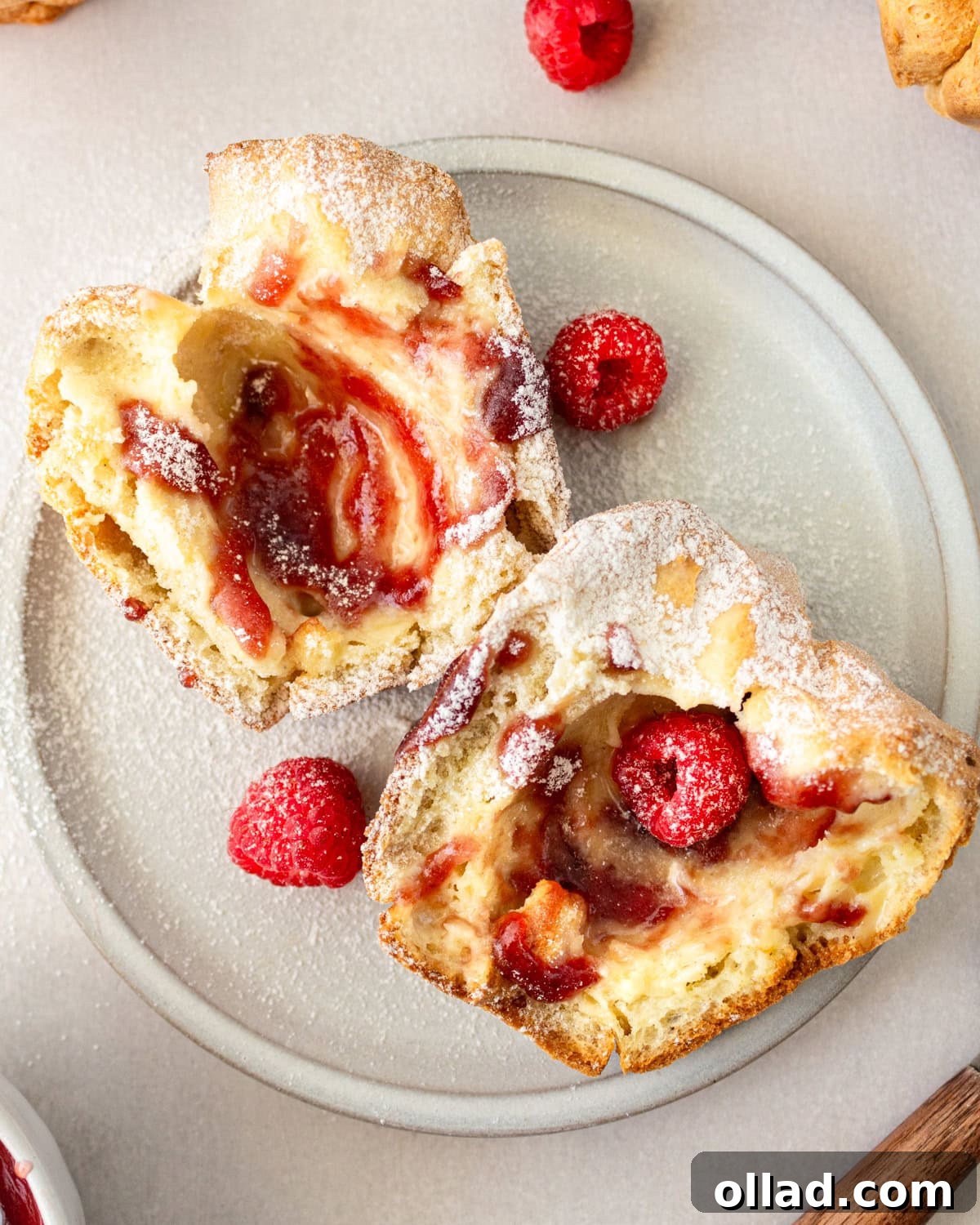 Overhead view of a gluten-free popover cut in half, revealing its airy interior, garnished with fresh raspberries and a dusting of powdered sugar on a white plate.