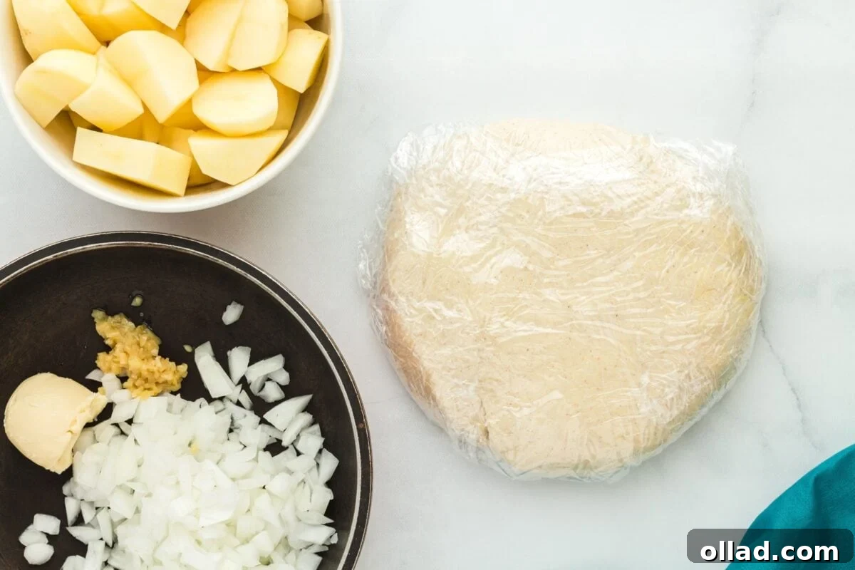 A disc of pierogi dough, tightly wrapped in plastic wrap, next to a black bowl of chopped onions and a white bowl of cubed potatoes, all ready for the next step.