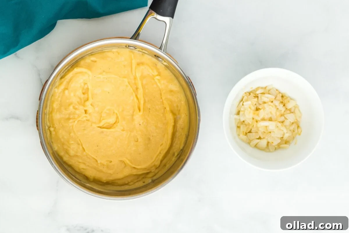 Overhead shot of rich, creamy mashed cheesy potatoes in a pot, with a white bowl of sautéed onions beside it, prepared for mixing.