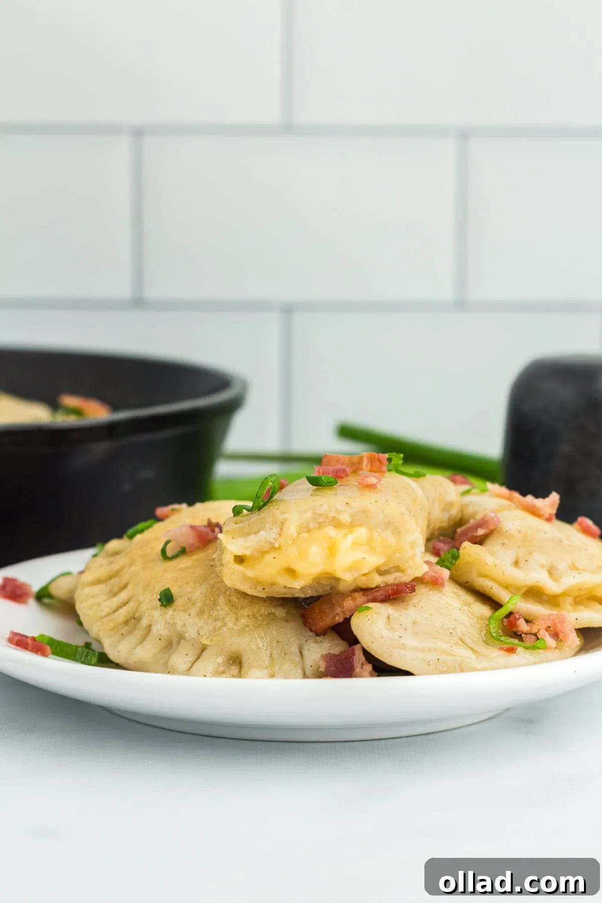 Delectable Gluten-Free Pierogi 5 Side view of a serving of gluten-free pierogi on a white plate, with a cast iron pan filled with more pierogi in the soft-focus background.