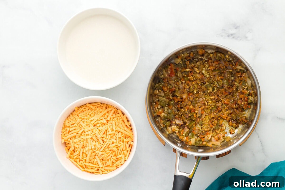 Sautéed onions, bell pepper, jalapeños, and garlic in a stainless steel pot, with a bowl of shredded vegan cheese and a can of evaporated coconut milk nearby, ready for the next steps.