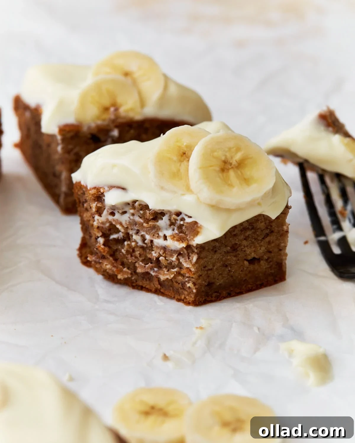 Irresistible Gluten-Free Banana Cake 2 A piece of gluten-free banana cake topped with cream cheese frosting and banana slices with another cake slice behind
