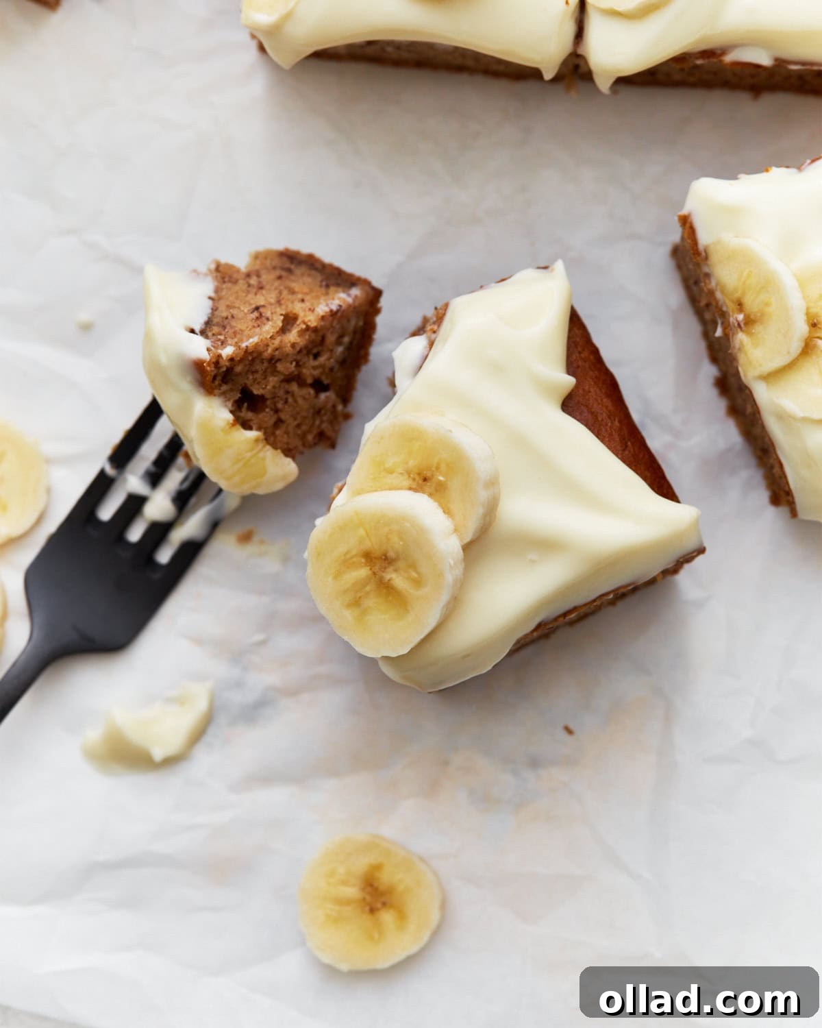 Irresistible Gluten-Free Banana Cake 13 Gluten-free banana cake slices topped with creamy frosting and fresh banana slices, placed on parchment paper with a fork holding a piece of the cake.