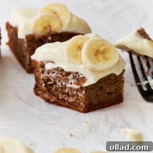 A close-up of a slice of gluten-free banana cake topped with creamy frosting and fresh banana slices, resting on parchment paper with a fork in the background