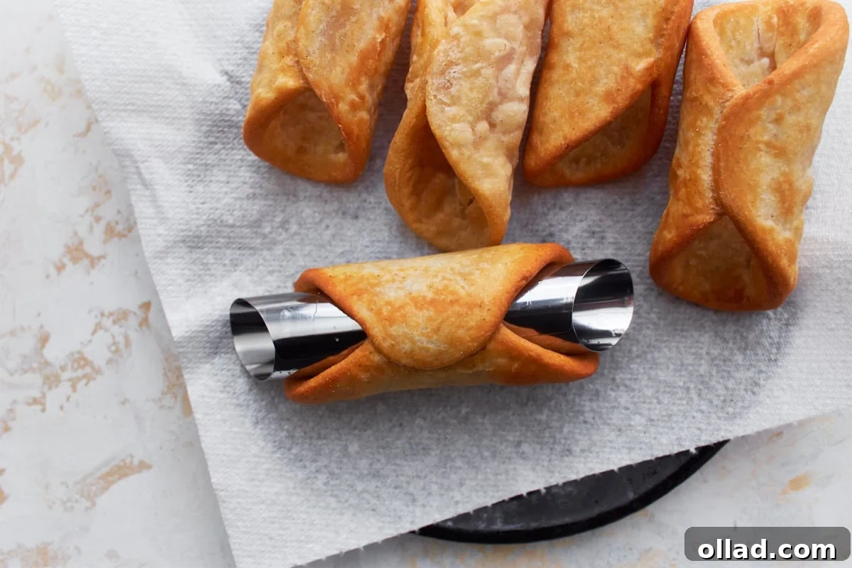 Golden-brown cannoli shells cooling on a paper towel-lined surface, with one shell still wrapped around a shiny metal mold. The shells have a crisp, bubbly texture.