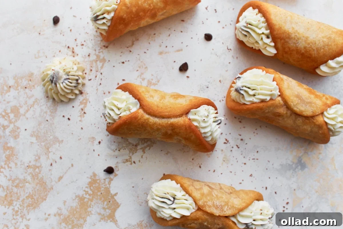 Overhead view of gluten-free cannoli arranged on a textured light surface, filled with ricotta cream piped at both ends. Chocolate chips and a dollop of cream are scattered around for decoration.