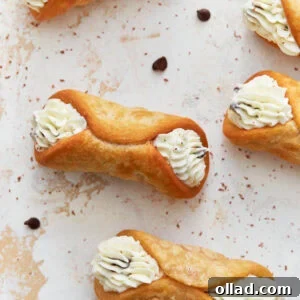 A close-up image of crispy, golden gluten-free cannoli shells filled with piped creamy ricotta filling, garnished with chocolate chips and dusted with cocoa powder on a light-textured surface.