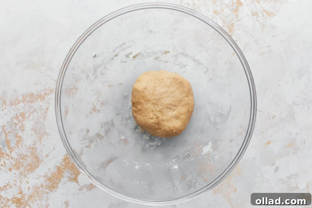 A ball of smooth gluten-free cannoli dough resting in the center of a clear glass mixing bowl, placed on a light-colored, textured surface 