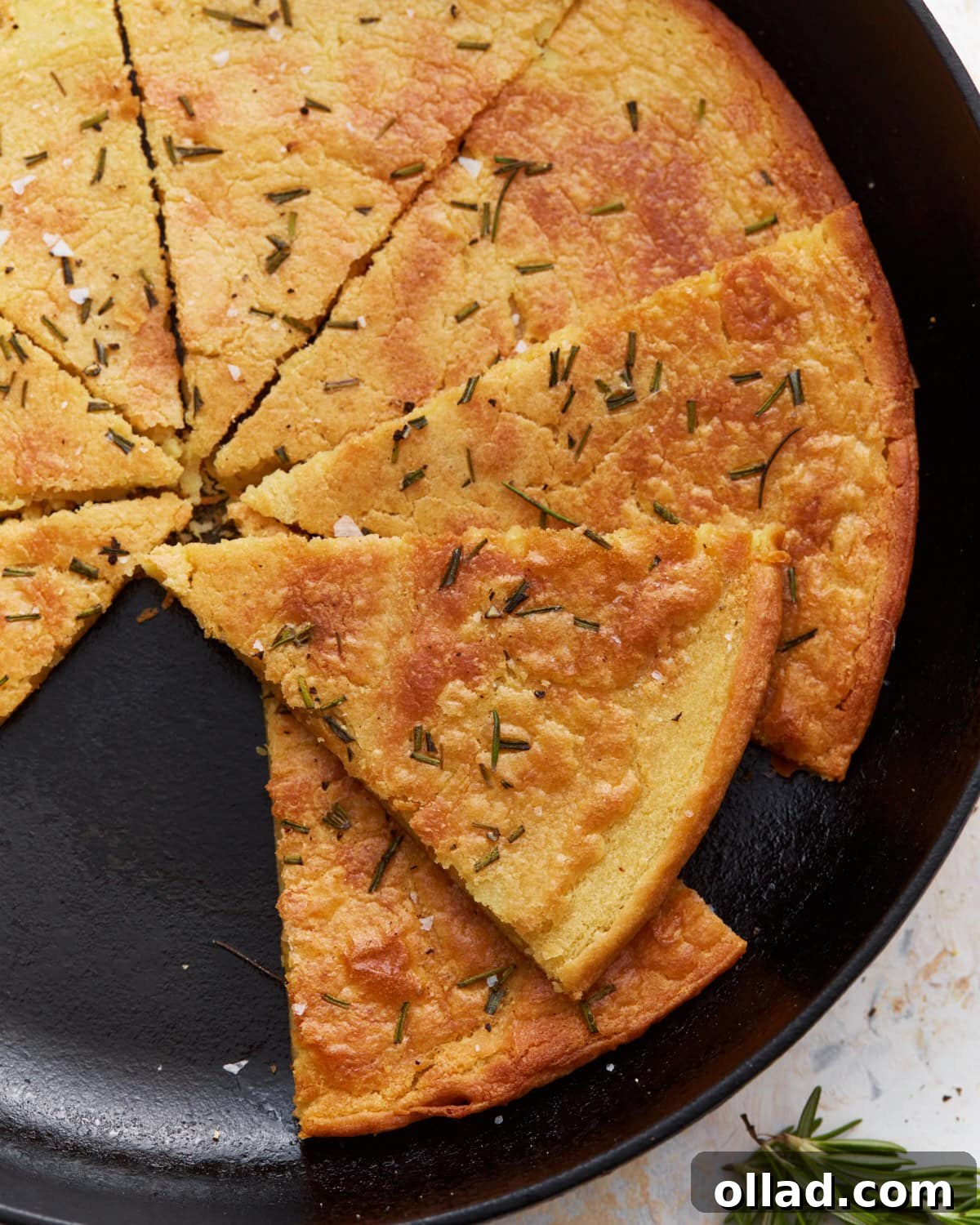 A sliced farinata (chickpea flatbread) in a cast iron skillet, garnished with fresh rosemary and flaky sea salt, showcasing its golden brown, crispy edges and soft interior.