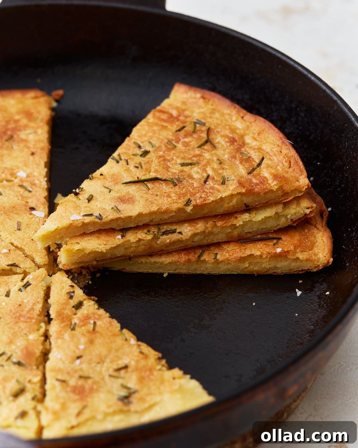 A side view of golden-brown farinata slices, stacked neatly in a cast iron skillet, highlighting their texture and light, crispy crust, garnished with rosemary and flaky salt.