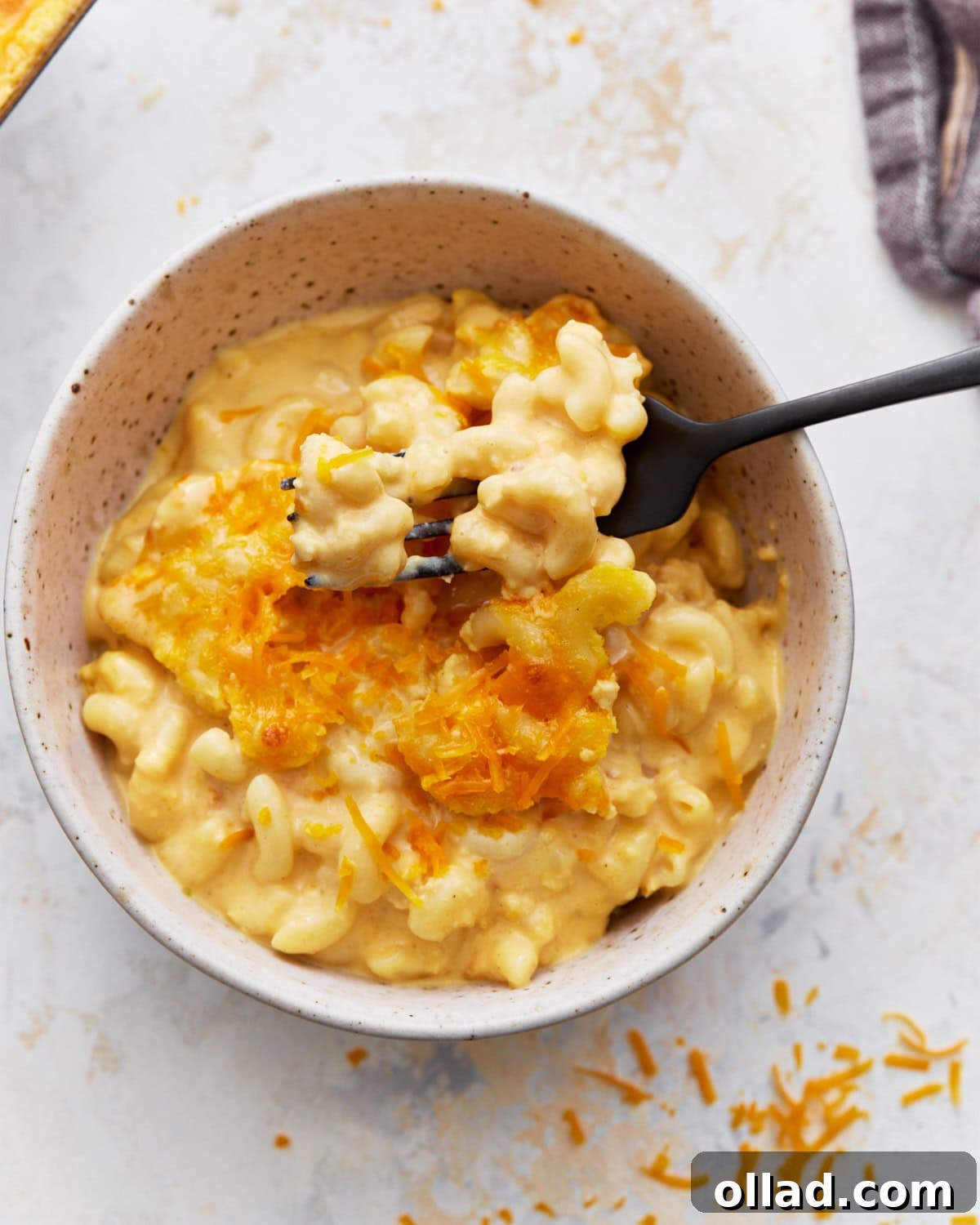 A fork lifting out a bite of baked gluten-free macaroni and cheese topped with shredded orange cheese from a white bowl, showing its creamy texture.