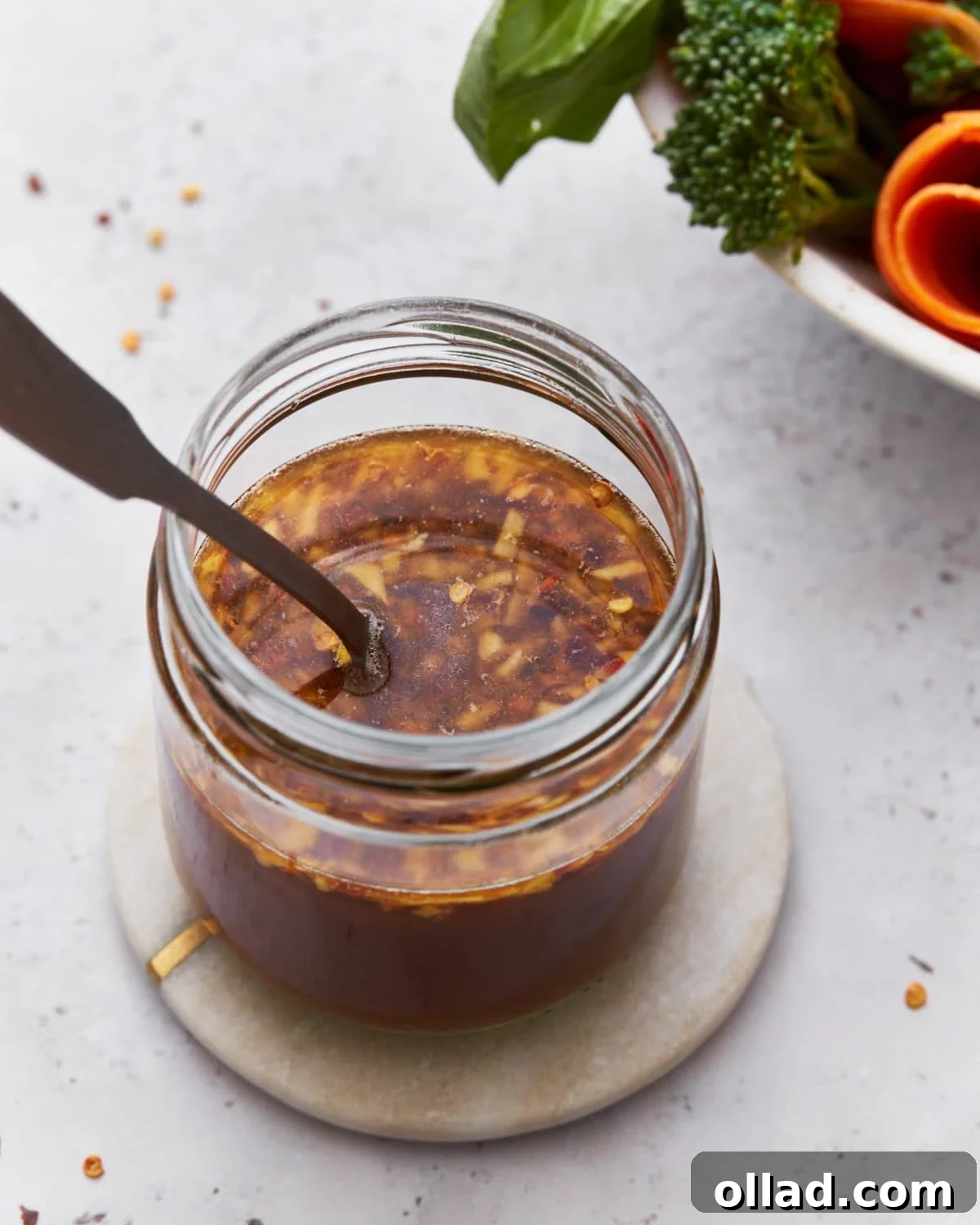 A close-up of a jar filled with gluten-free stir-fry sauce, featuring visible bits of garlic and red chili flakes. The jar sits on a small round coaster, with vibrant fresh broccoli and carrots blurred in the background.