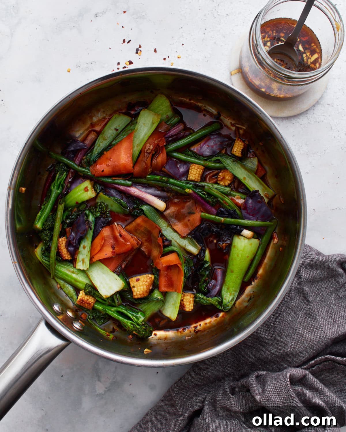 A sauté pan filled with colorful stir-fried vegetables, including bok choy, baby corn, carrots, and purple cabbage, tossed in a glossy gluten-free stir-fry sauce. A jar of the sauce sits nearby.