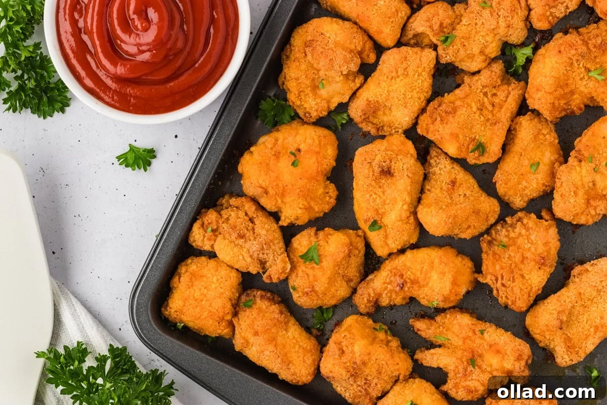 Overhead close-up of baked chicken nuggets on a sheet pan with a bowl of ketchup and sprigs of parsley nearby.