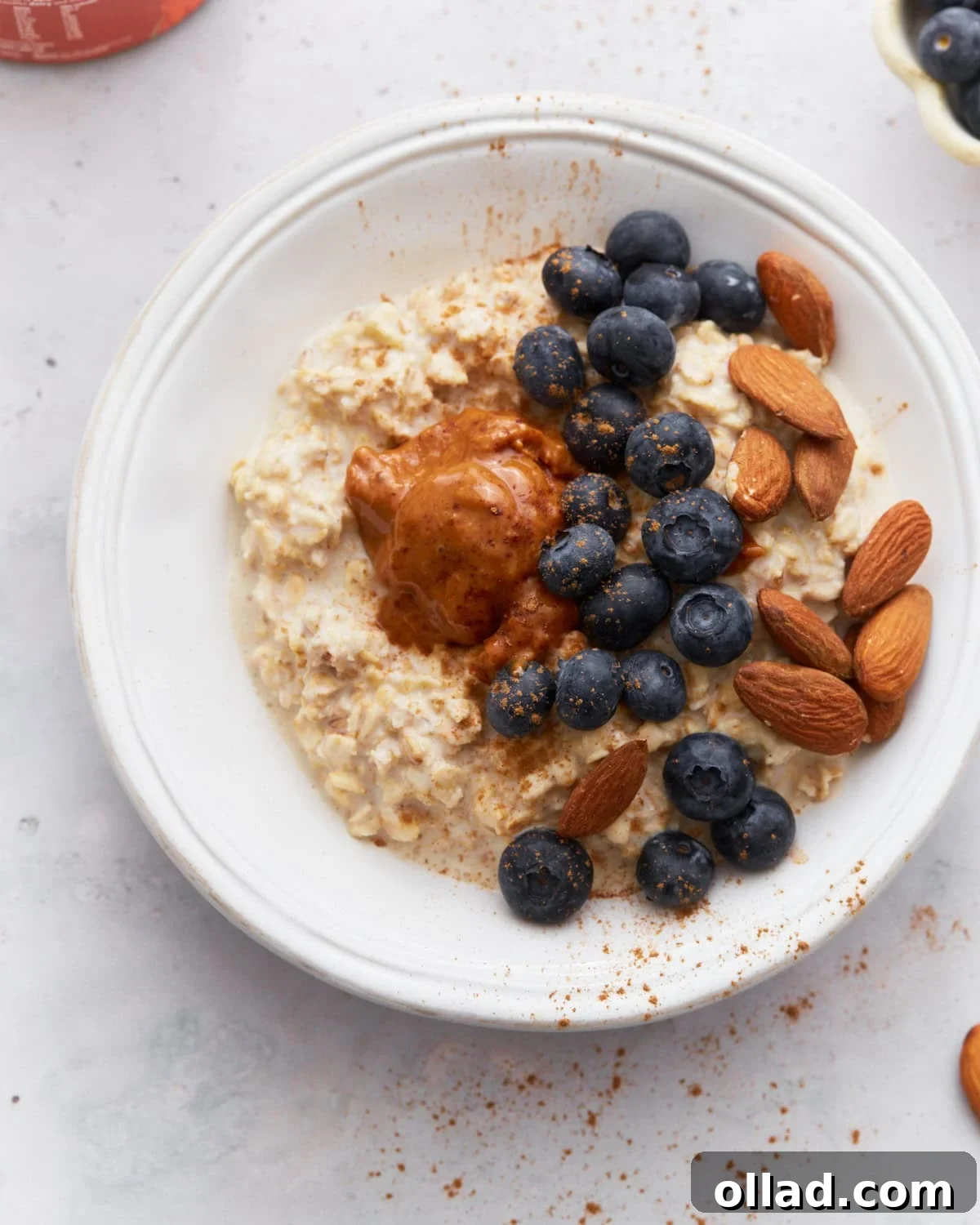 A bowl of creamy gluten-free overnight oats topped with fresh blueberries, whole almonds, and a dollop of almond butter, sprinkled with cinnamon, on a light surface.