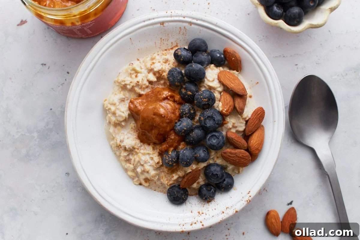 A top view of a beautifully arranged bowl of gluten-free overnight oats with blueberries, almonds, almond butter, and a sprinkle of cinnamon, alongside a spoon and additional toppings scattered around.