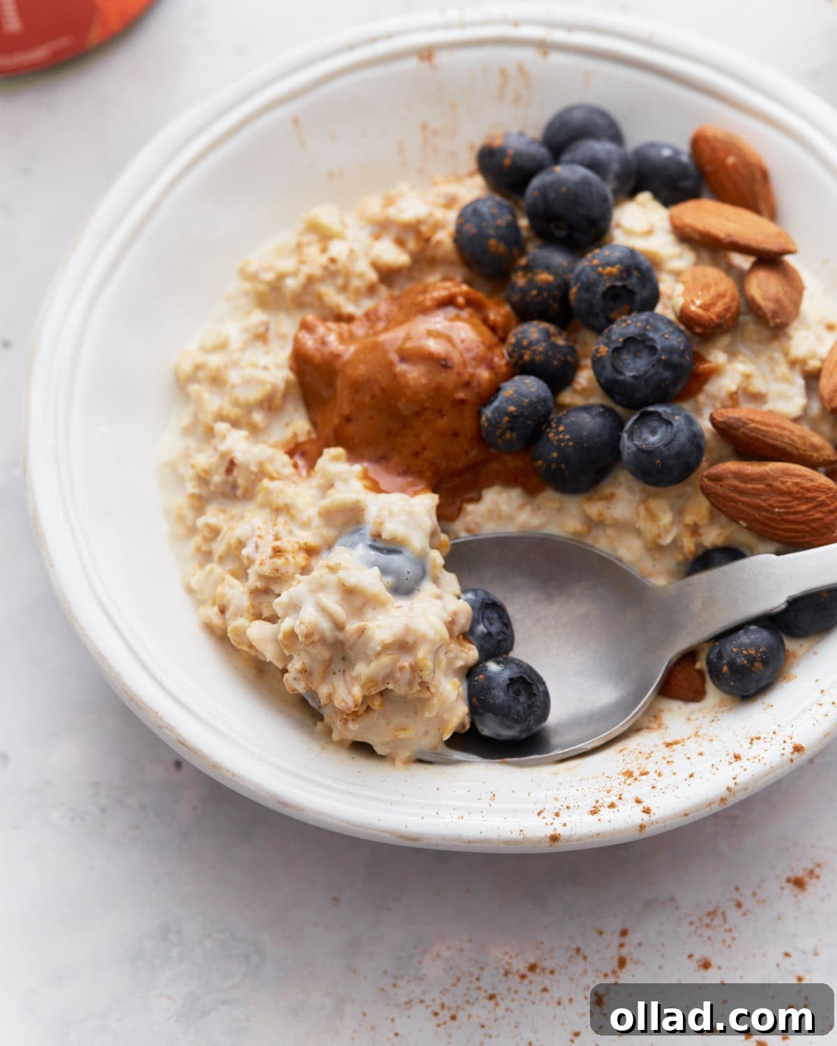 A close-up of gluten-free overnight oats with toppings, including blueberries, almonds, and almond butter, with a spoon scooping up a bite.