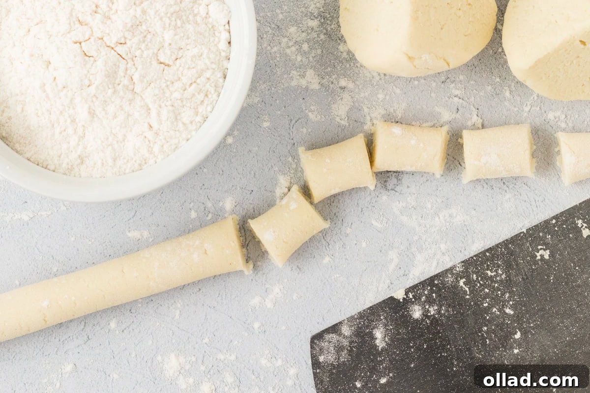 Pillowy Homemade Cauliflower Gnocchi 11 A close-up view of a perfectly rolled-out log of cauliflower gnocchi dough, precisely cut into small, bite-sized pieces using a sharp knife. The floured surface, a bowl of flour, and a dough scraper are visible, creating a dynamic scene of preparation.