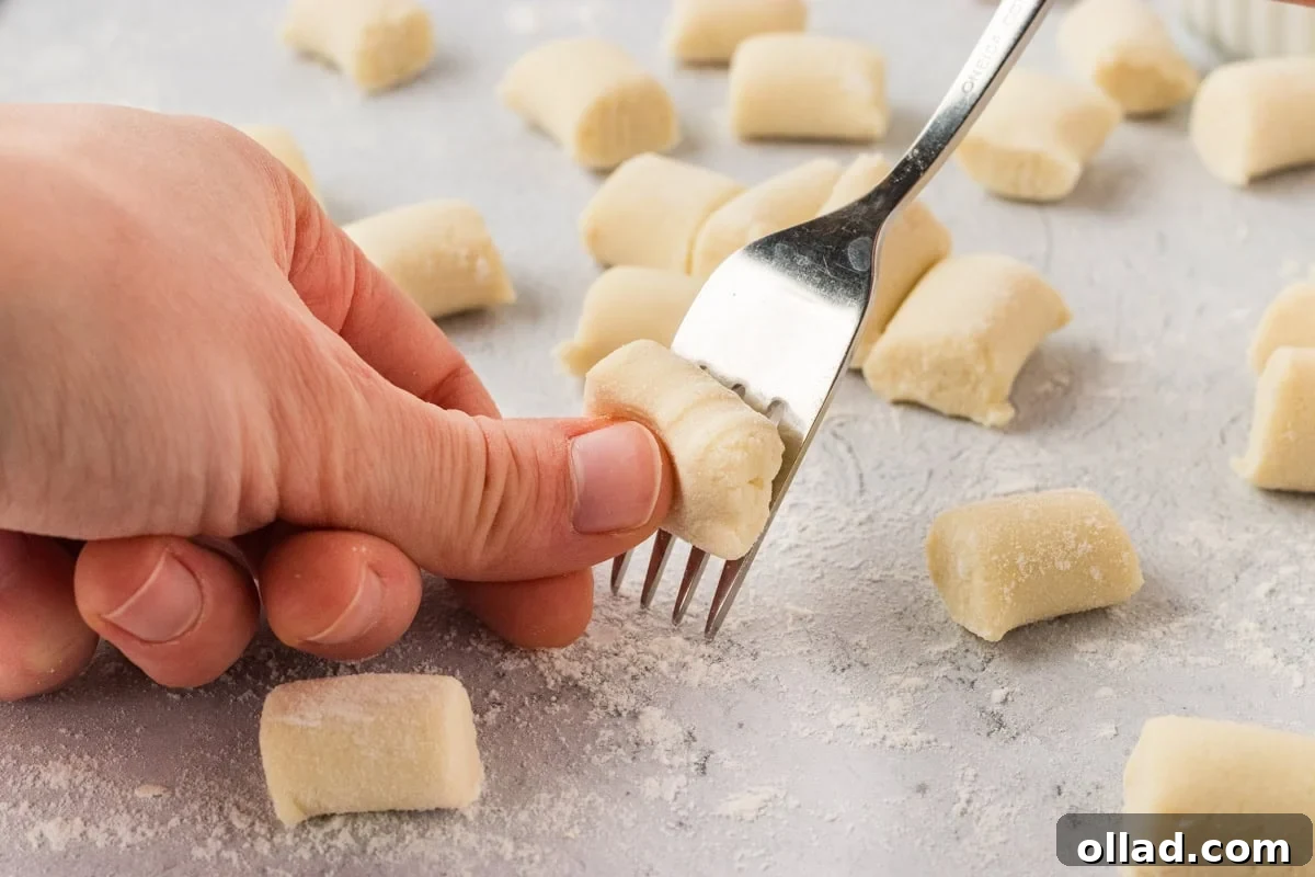 Pillowy Homemade Cauliflower Gnocchi 12 A close-up of a hand gently pressing a fork onto a small, individual piece of cauliflower gnocchi dough, creating the classic ridges that help sauce adhere. Several other pieces of shaped dough rest on a lightly floured surface, awaiting their turn.
