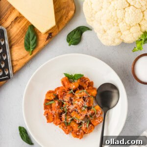 Pillowy Homemade Cauliflower Gnocchi 3 A white plate filled with golden brown cauliflower gnocchi covered in tomato sauce, garnished with fresh basil and grated cheese. A black spoon rests on the plate, surrounded by ingredients like Parmesan, fresh basil, and a head of cauliflower.