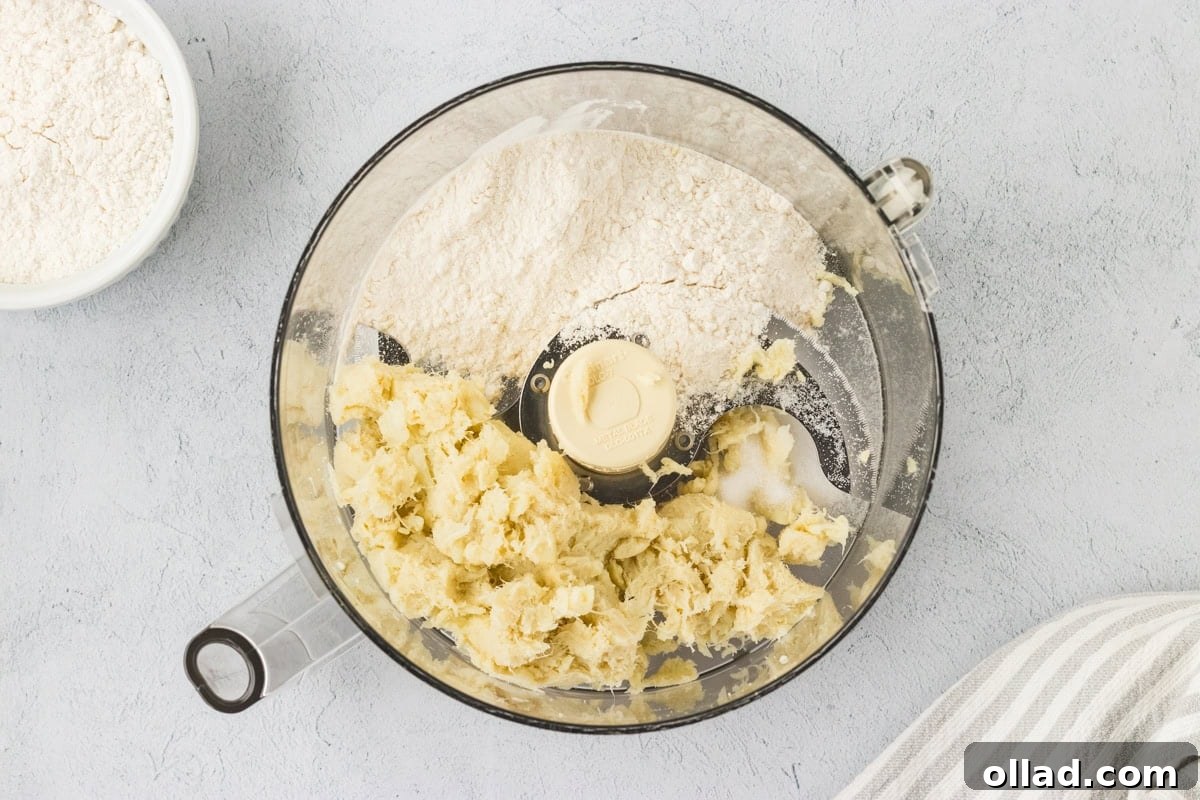 Pillowy Homemade Cauliflower Gnocchi 7 A bird's-eye view of a food processor bowl, containing a creamy mixture of well-drained mashed cauliflower, gluten-free all-purpose flour, and salt, perfectly blended into a soft dough. A small bowl of extra flour is visible nearby for dusting.