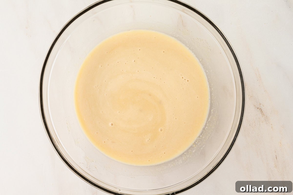 A glass mixing bowl filled with combined wet ingredients for the donut batter, showing a smooth and slightly frothy texture.