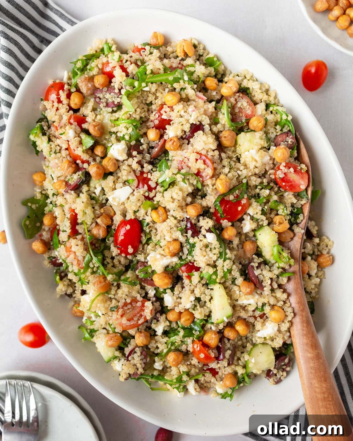 Mediterranean Quinoa Bowl 2 A white oval dish filled with Greek quinoa salad, featuring fluffy quinoa, cherry tomatoes, cucumbers, Kalamata olives, arugula, feta cheese, and crispy roasted chickpeas. A wooden spoon rests in the dish.
