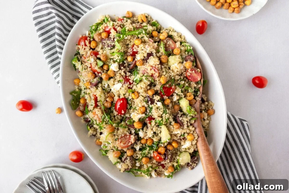 Mediterranean Quinoa Bowl 10 A fully assembled Greek quinoa salad served in a white oval dish, garnished with crispy roasted chickpeas and fresh herbs. A striped napkin and extra tomatoes surround the dish.