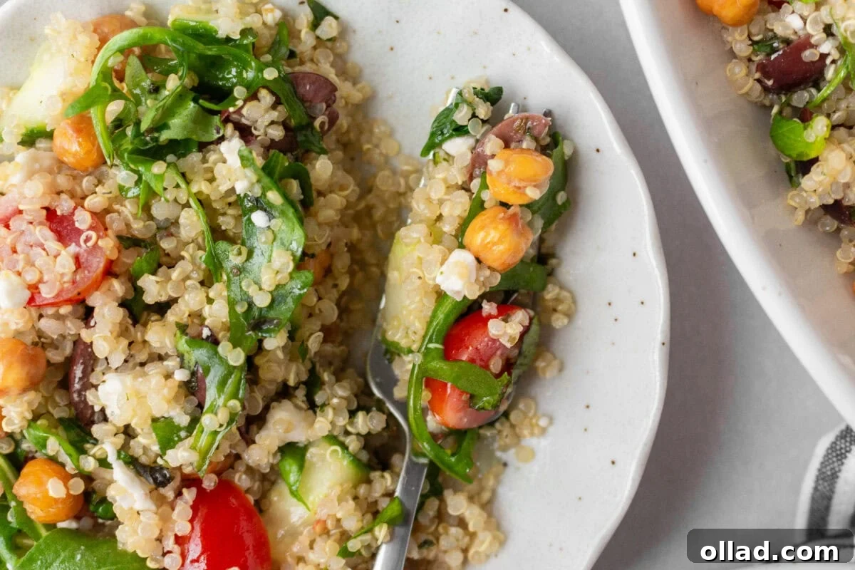 Mediterranean Quinoa Bowl 12 A close-up shot of a forkful of Greek quinoa salad, showcasing fluffy quinoa, arugula, cherry tomatoes, Kalamata olives, crispy roasted chickpeas, cucumbers, and crumbles of feta cheese on a white speckled plate. The vibrant ingredients highlight the fresh and flavorful textures of the dish.