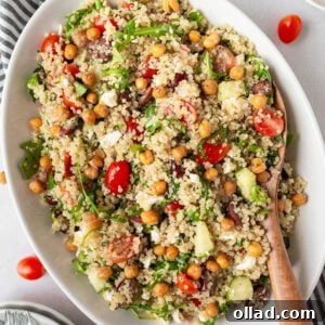 A white oval dish filled with Greek quinoa salad, featuring fluffy quinoa, cherry tomatoes, cucumbers, Kalamata olives, arugula, feta cheese, and crispy roasted chickpeas. A wooden spoon rests in the dish.