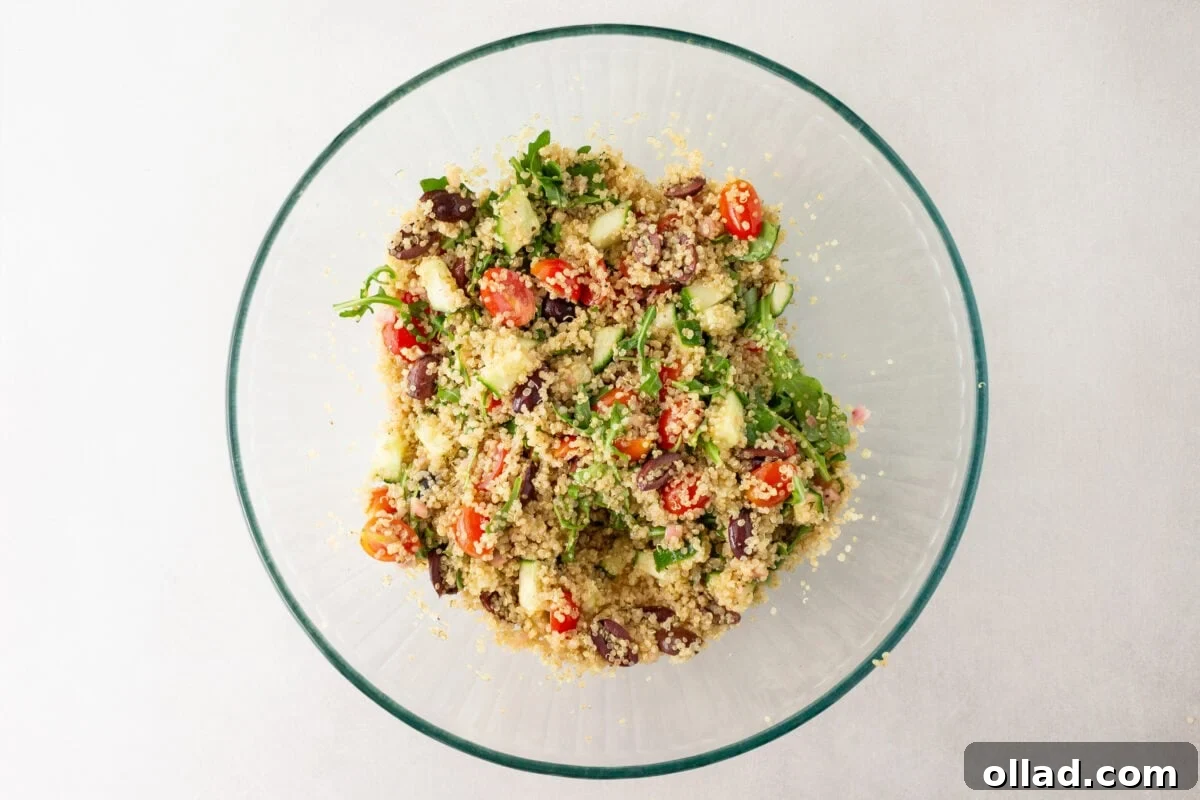 Mediterranean Quinoa Bowl 9 A large glass bowl filled with freshly mixed Greek quinoa salad, showcasing bright ingredients like cherry tomatoes, cucumbers, Kalamata olives, arugula, and feta cheese.