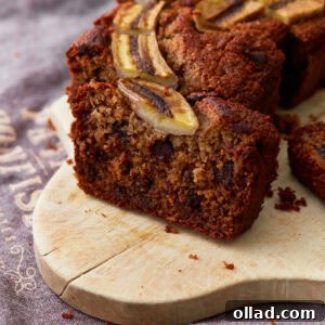 A close-up of a sliced loaf of banana oatmeal bread with dark chocolate chips and a caramelized banana topping, sitting on a wooden board.