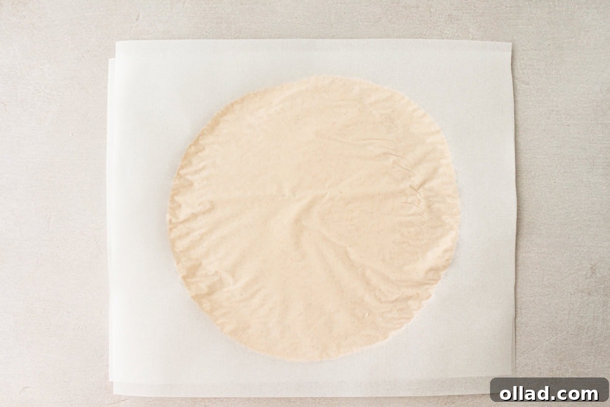 A skillfully rolled-out cassava tortilla rests perfectly between two sheets of parchment paper, showcasing its even thickness and readiness for cooking.
