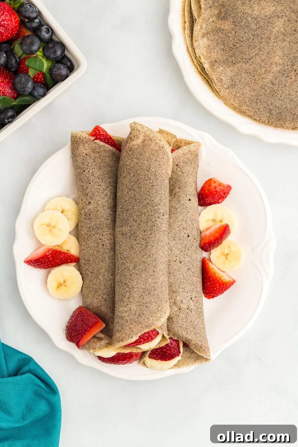 Buckwheat Galettes 2 Three rolled buckwheat crepes on a white plate, surrounded by fresh banana slices and halved strawberries.