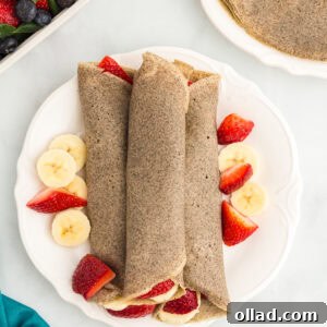 Three rolled buckwheat crepes on a white plate, surrounded by fresh banana slices and halved strawberries.