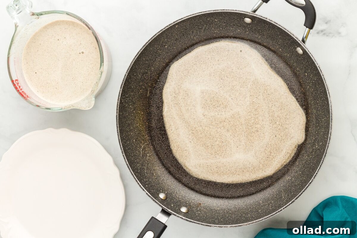 Buckwheat Galettes 7 A buckwheat crepe cooking in a skillet, forming a thin circular layer, with crepe batter in a measuring cup nearby.