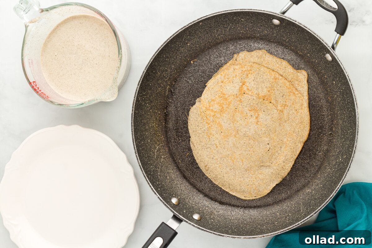 Buckwheat Galettes 8 A golden-brown buckwheat crepe cooked on one side in a skillet, with a measuring cup of batter and an empty plate beside it.