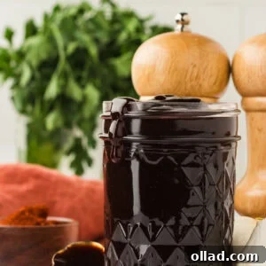 A glass jar filled with thick, dark homemade gluten-free BBQ sauce, with sauce dripping slightly over the rim. Fresh parsley, garlic, and wooden pepper mills are in the background, emphasizing the fresh, homemade quality of the sauce.