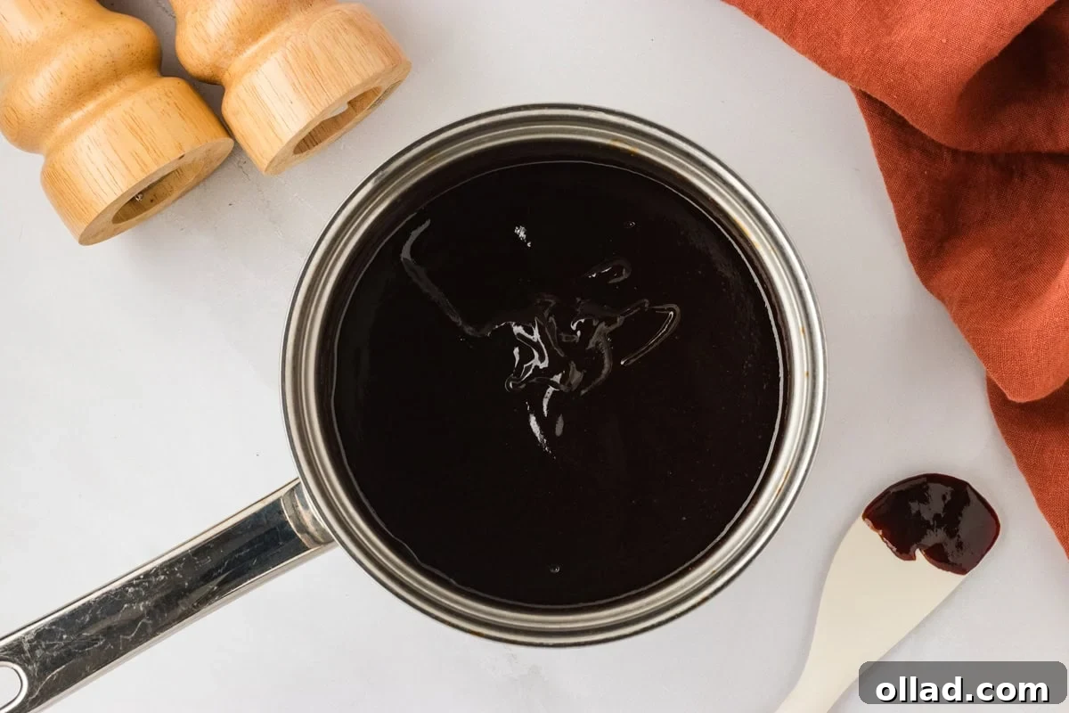 Crafted Gluten-Free BBQ Sauce 6 The homemade gluten-free BBQ sauce now fully mixed and visibly thickened, simmering gently in the saucepan, exhibiting a smooth, glossy, and rich dark red-brown finish. A clean white spatula with a small trace of the sauce rests on the counter beside the pot, indicating the delicious process.